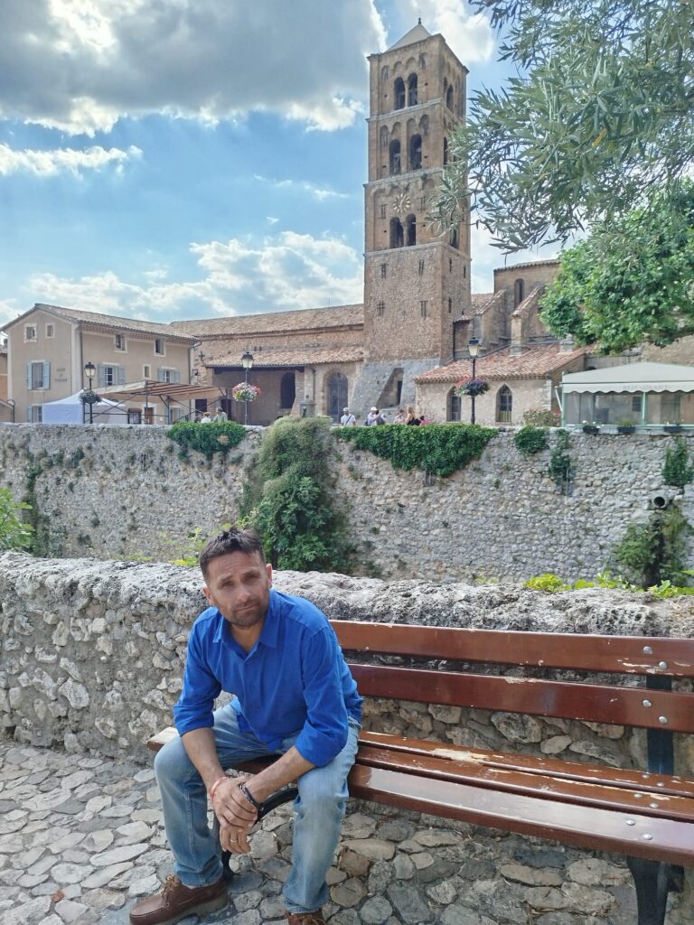 Daryl assis sur un banc devant l'église Notre-Dame-de-l'Assomption de Moustiers-Sainte-Marie, en Provence, pendant le tournage du clip Enfant de la Provence.